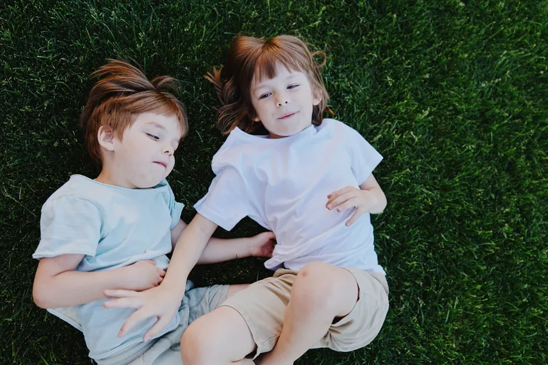 Two children playing on green grass, enjoying a sunny day, showcasing friendship and laughter with carefree expressions and casual clothing.