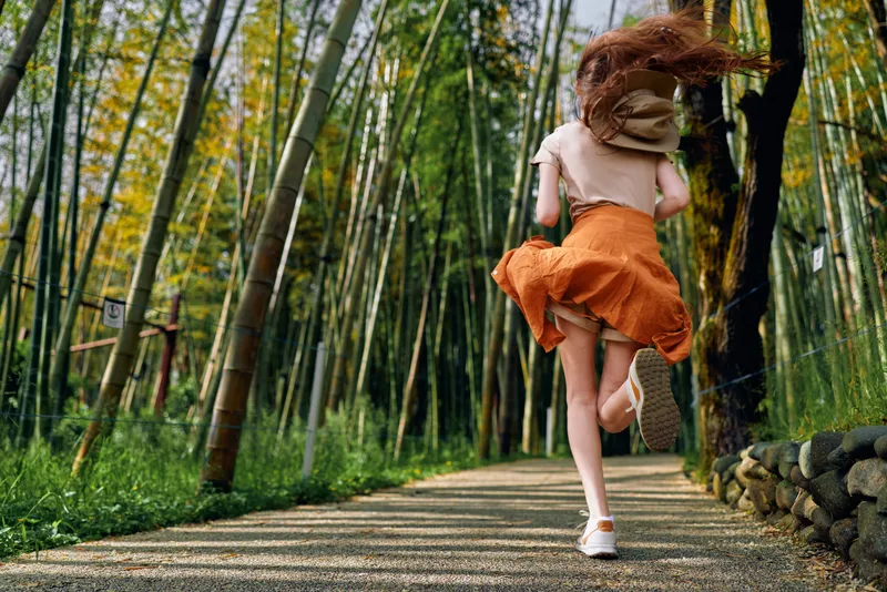Girl running on a gravel path through a sunlit bamboo forest wearing an orange skirt and white sneakers, redhair flowing, energetic outdoor motion and scenic trail perspective.