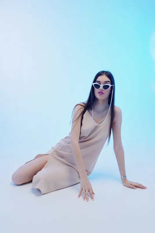 Young woman in a beige dress and stylish sunglasses poses on the floor against a soft blue background. Concept of woman beauty fashion with modern elegance and minimalism.