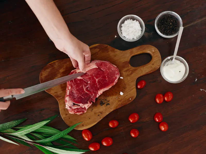 Женщина с ножом в руке режет свежее мясо стейка для жарки на кухне, на столе соль, перец и другие специи, красные помидоры черри и зелень, готовит ужин в домашней кухне
