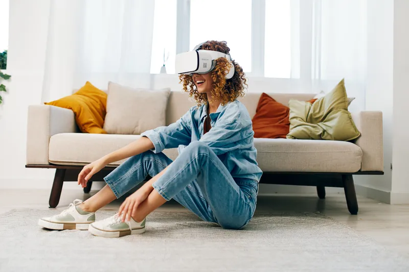 Улыбающаяся женщина наслаждается игрой в виртуальной реальности с VR-очками на диване в современной гостиной