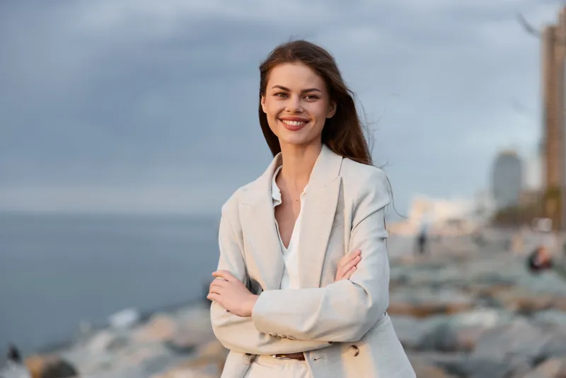 Confident businesswoman smiling outdoors near the shore during golden hour, dressed in a stylish light-colored suit, embodying professionalism and elegance.