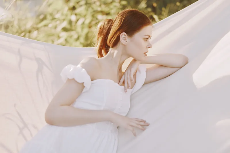 A serene young woman in a white dress relaxes in a hammock, surrounded by soft greenery and warm sunlight, embodying tranquility and summer vibes