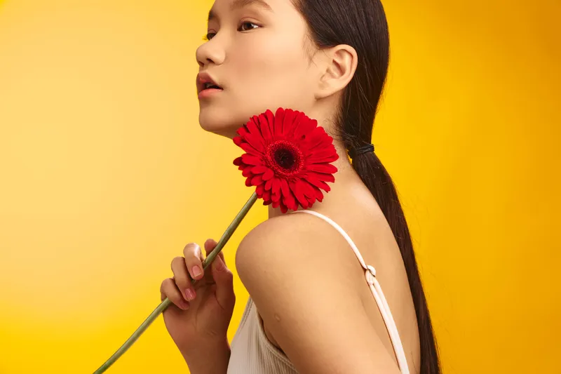 asian woman is framed in a bright studio setting, holding a red flower near her shoulder. she gazes softly away, exuding calm confidence and artistic nuance through clean lighting and minimal