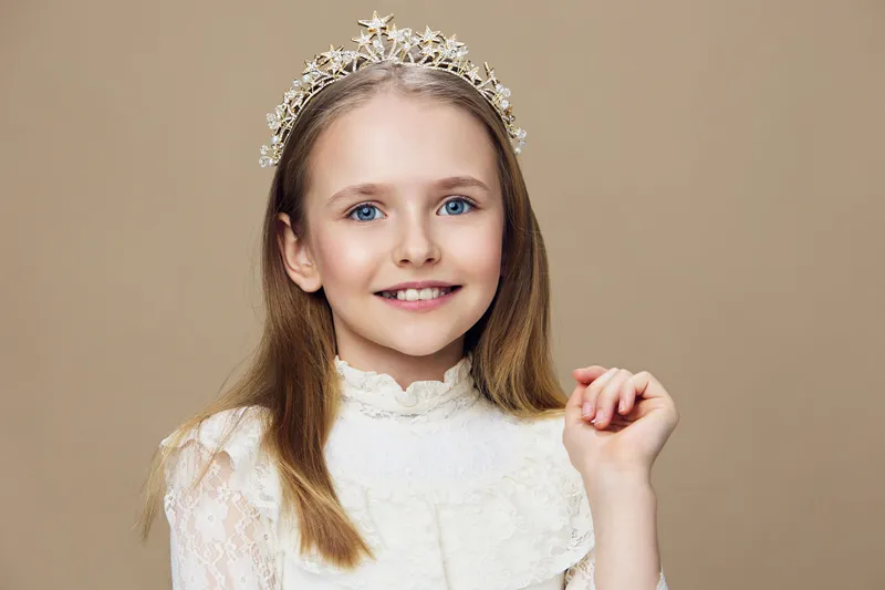 Smiling girl in a white royal dress with a crown, showcasing joy and elegance against a beige background, perfect for children s events or portraits