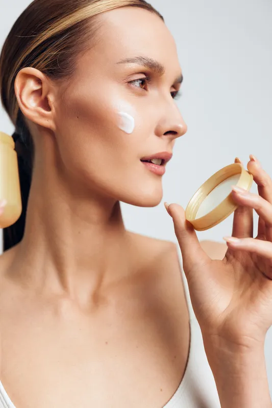 Young woman applies moisturizer from a small jar to her cheek in a calm skincare moment, soft lighting highlighting glowing skin and a clean minimal background, natural textures and serene expression.