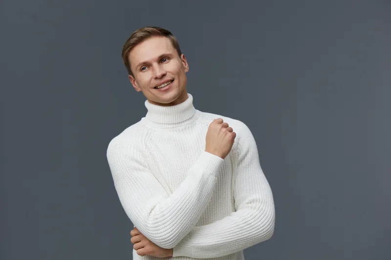 Smiling man wearing a cozy white turtleneck sweater posing with a friendly expression against a plain gray background for a lifestyle portrait emphasizing warmth and comfort. Concept: happy people.