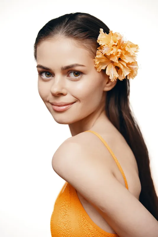 Портрет улыбающейся молодой женщины, украшенной оранжевым цветком и одетой в яркий оранжевый наряд, излучающей радость и летнюю эстетику на светлом фоне