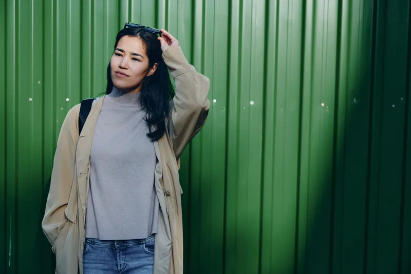 A woman in a gray sweater and beige coat stands beside a green wall, casual urban fashion and relaxed mood, showcasing contemporary street style and everyday wear