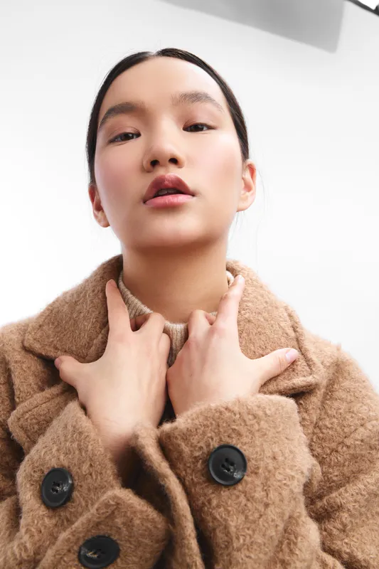 asian woman models a warm textured coat, posing in a clean studio. she gazes softly at the camera, exuding calm confidence and contemporary style.