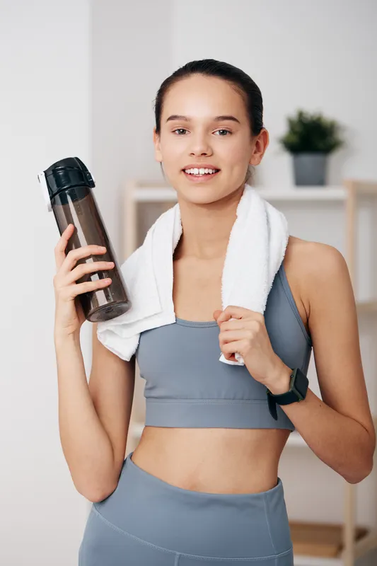 Woman fitness sport portrait of young woman after workout, wearing sports bra and leggings, holding water bottle with towel over shoulder, smartwatch visible, bright indoor home gym