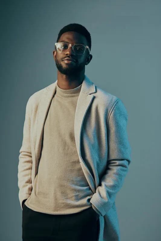 Stylish young Black man in a light sweater and coat, exuding confidence against a blue green background, perfect for fashion and lifestyle concepts