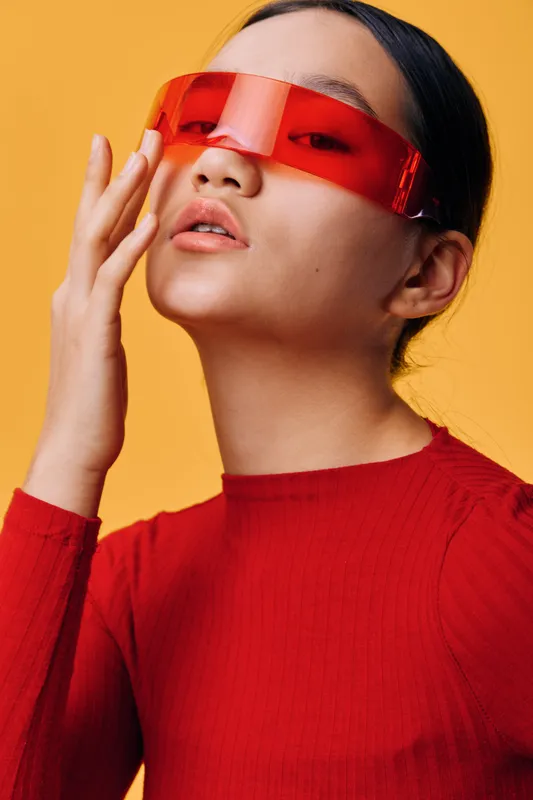 Woman wearing futuristic red sunglasses and red ribbed sweater posing with hand near face against orange background. Closeup portrait with stylish eyewear and modern fashion expression.