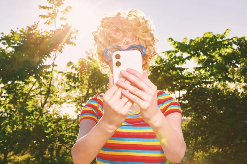 A young person with curly hair wearing sunglasses and a striped shirt takes a selfie outdoors, bright light and lens flare creating a playful, retro mood. Concept for youth, outdoor photography,