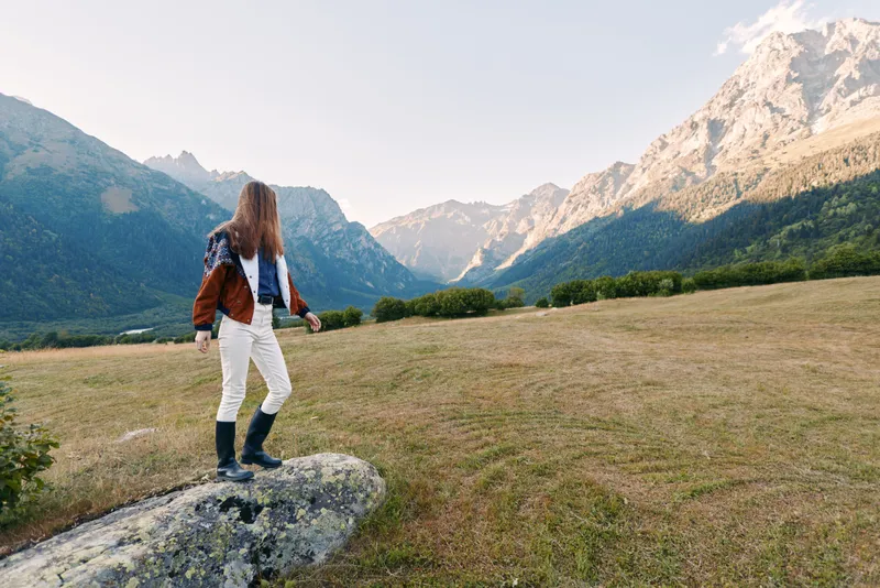 Woman standing on a rock in a mountain meadow, hiking through nature landscape as a solo traveler outdoors, wearing boots and jacket, looking toward distant peaks.