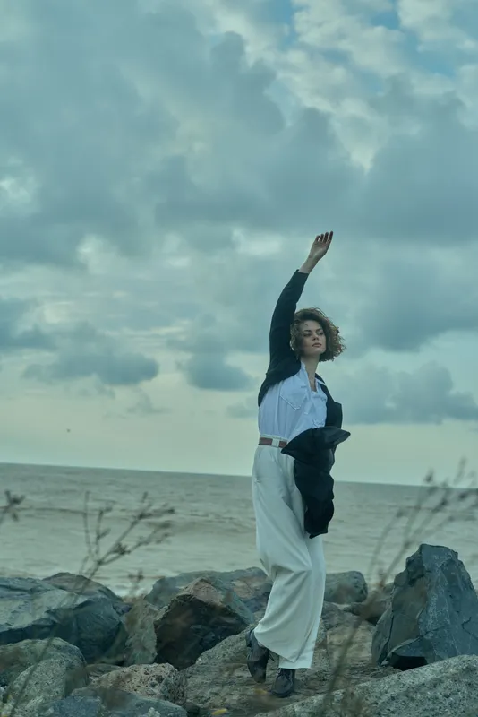 Woman stands on rocky coast by the sea with a raised arm, wind in hair, wearing a white dress and dark jacket, capturing a moody outdoor moment against the expansive horizon