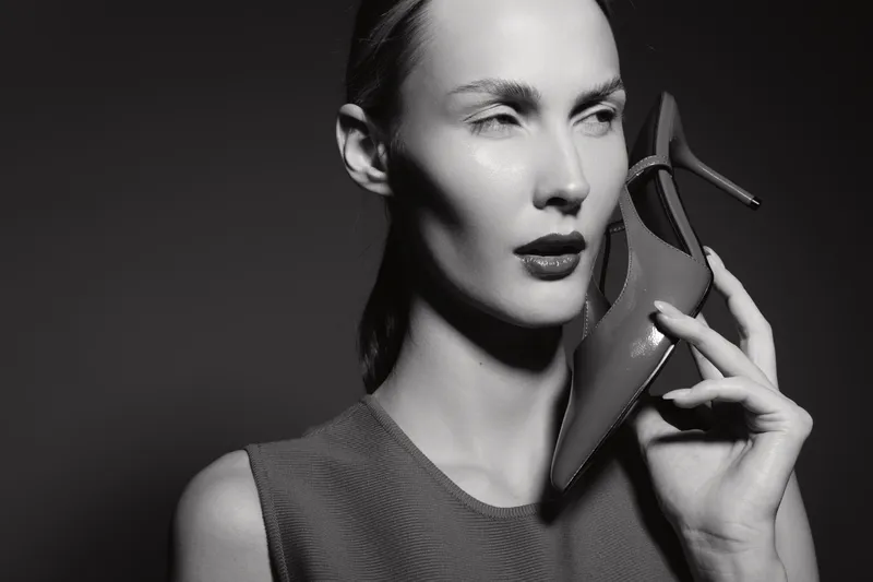 Monochrome portrait of a fair skinned caucasian woman holding a high heel shoe near her face. Stylish female model poses with a serious expression against a dark background.