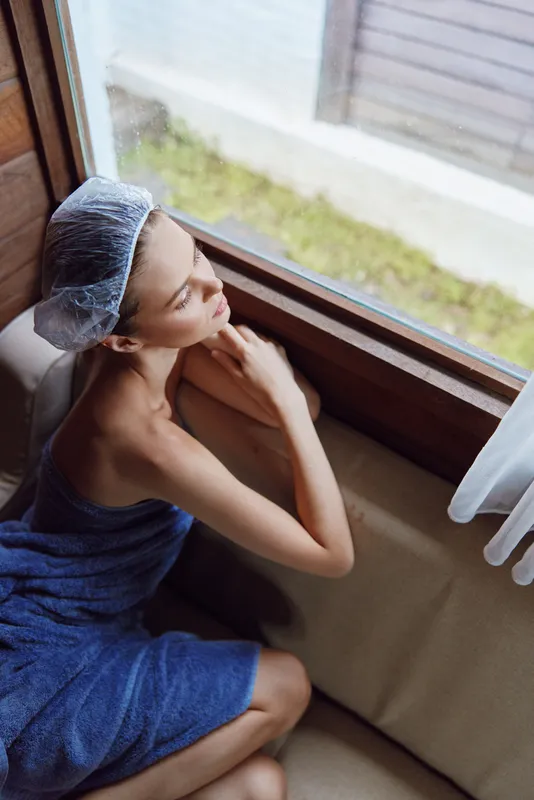 woman in bath towel and shower cap sitting by window, relaxed and thoughtful, soft natural light, indoor self-care moment, calm and peaceful atmosphere