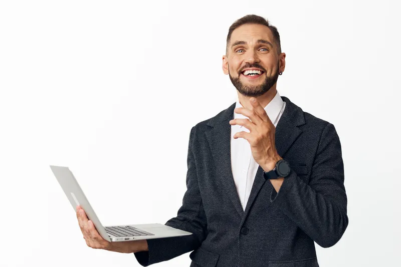 A professional man in a smart blazer smiles warmly while holding a laptop, standing against an isolated white background to convey confident business presence and positive emotion.
