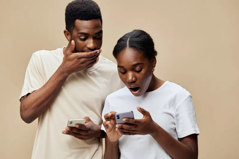 Shocked expressions of a young Black couple looking at their smartphones in casual attire against a light beige background, capturing a moment of surprise