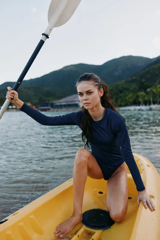Young female athlete kneels in a yellow kayak, gripping a paddle, prepared to push off into calm water, with distant mountains framing the scene and clear sky above.