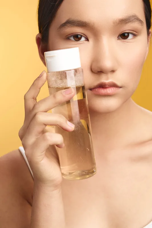 asian woman holds a clear bottle of liquid skincare against a warm yellow backdrop, creating a clean beauty mood and inviting routine focused on selfcare and glow.
