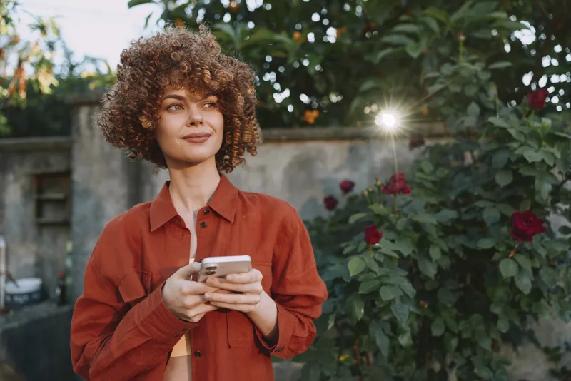 Cheerful woman outdoors, orange shirt, holding a smartphone, garden background bathed in sunlight, relaxed pose, casual lifestyle, natural colors, candid vibe, smiling