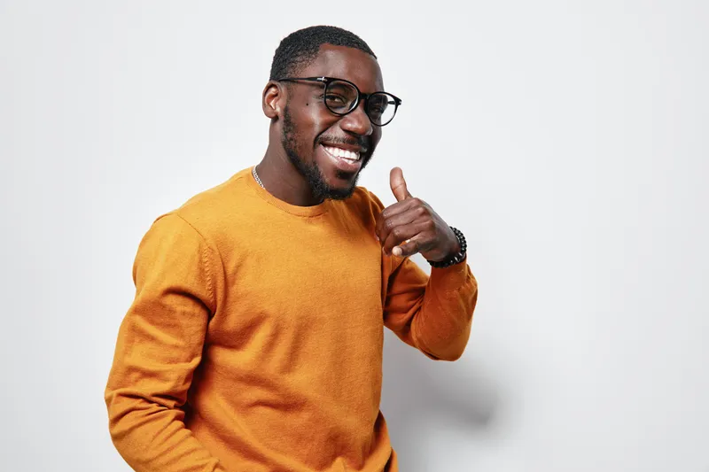 African American man with dark skin wearing glasses and orange sweater smiling and making call me gesture on white background. Casual style and positive expression.