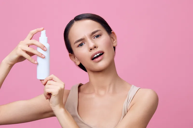 Young woman with straight hair in a beige top presenting a hair care product against a pink background, showcasing emotions of surprise and curiosity
