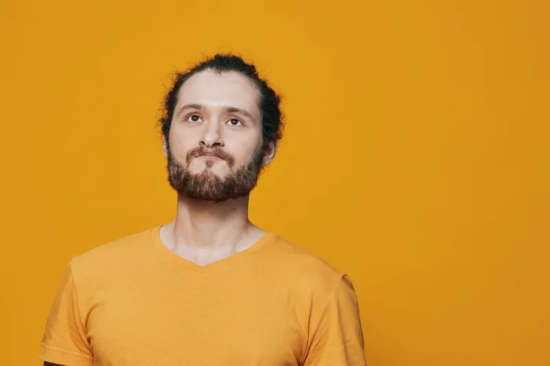 Male model with beard and bun hairstyle smiling against a bright yellow background, wearing a casual orange T shirt, evoking a positive and cheerful mood