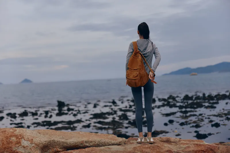 Young woman in casual grey hoodie and backpack gazing at the ocean, evoking a sense of adventure and tranquility in a coastal landscape