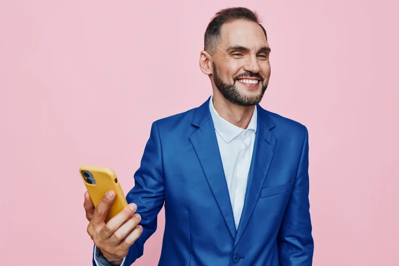 A confident business professional smiles while looking at a phone against a solid pink background, conveying positive emotion and polished style.