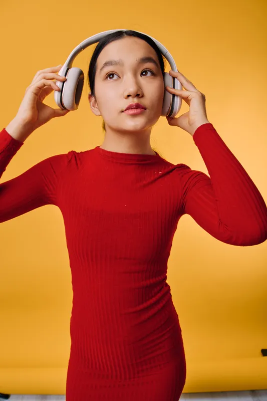 Young woman in red dress adjusting wireless headphones with thoughtful expression on yellow background. Female, fashion, technology, and music lifestyle concept in studio.
