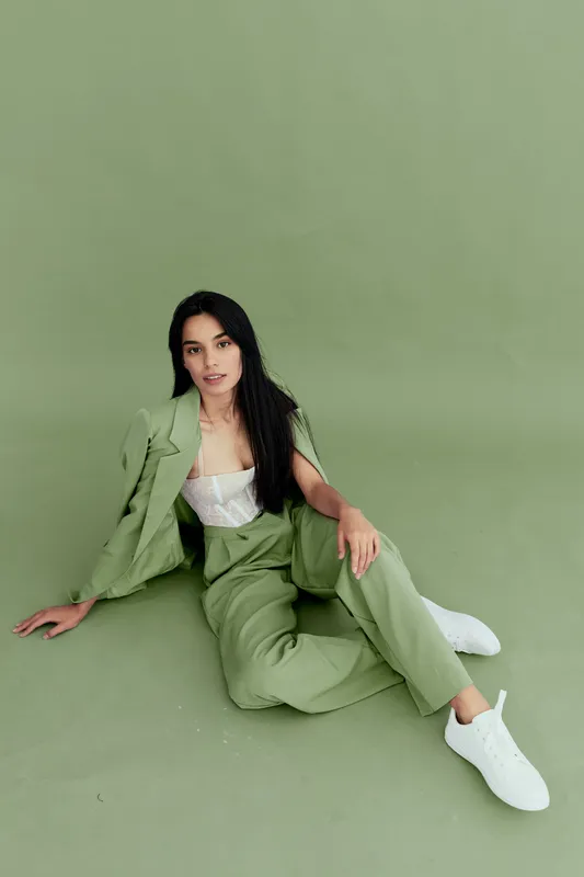 Young woman in green pantsuit sitting on floor, wearing white top and sneakers against matching green background, casual style, relaxed pose, studio setting.