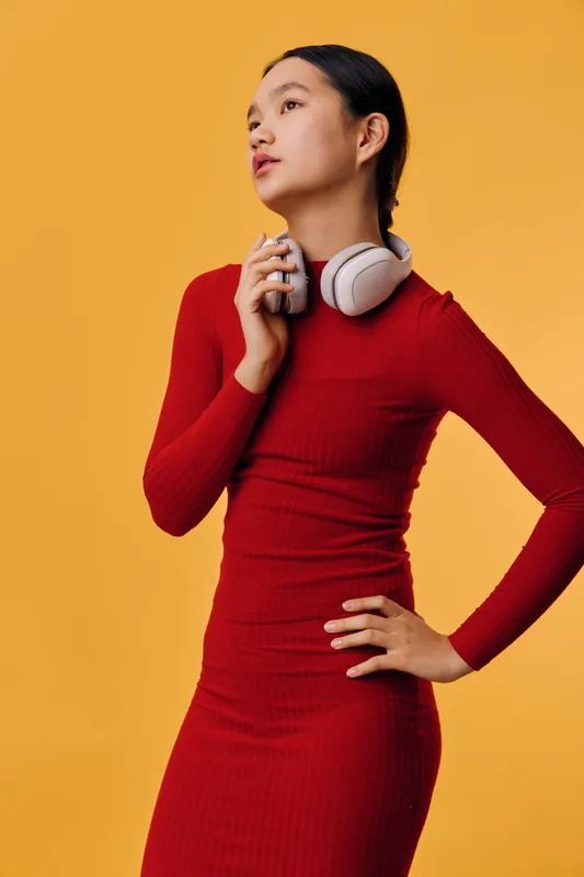 Young woman in red dress holding white headphones posing with hand on hip looking away on yellow background. Fashion model portrait with casual style and confident pose.