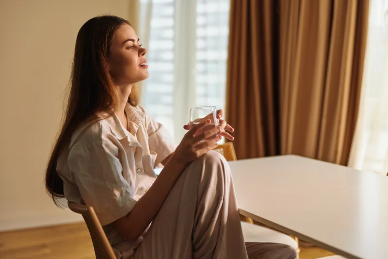 Woman sitting at home by a table holding a glass, relaxed and smiling in morning sunlight through window curtains, casual loungewear and peaceful domestic routine.