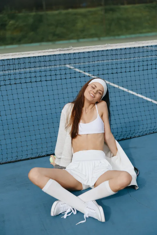 Smiling athletic woman relaxing on a tennis court in sporty white outfit, showcasing confidence and joy, emphasizing healthy lifestyle and sports enthusiasm