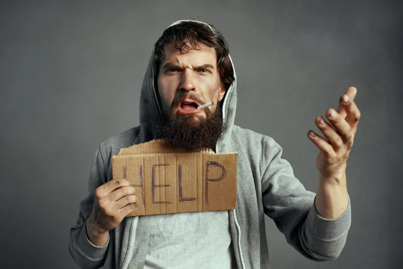 Бездомный мужчина с бородой держит табличку с надписью «Help» на улице