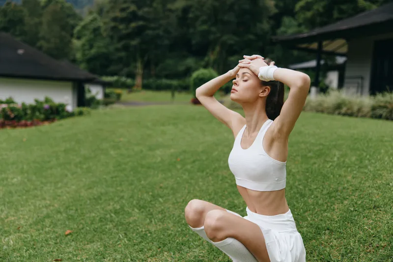 Young model with a serene expression in white athletic wear, practicing yoga in a lush green outdoor setting, representing fitness and mindfulness