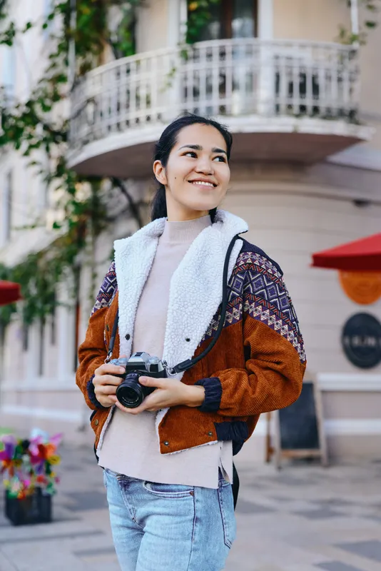 Camera in hand, smiling woman stands outdoors in a cozy brown jacket, casual street style, urban setting, ready for lifestyle photography and travel shoots.