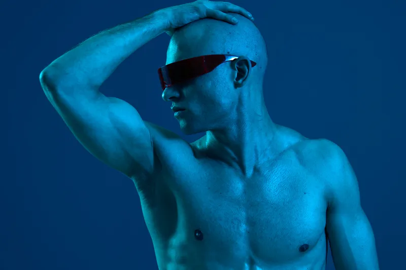 Confident man in cool sunglasses poses against a blue background, showcasing an artistic touch with dramatic lighting. This striking image combines fashion and emotion uniquely.