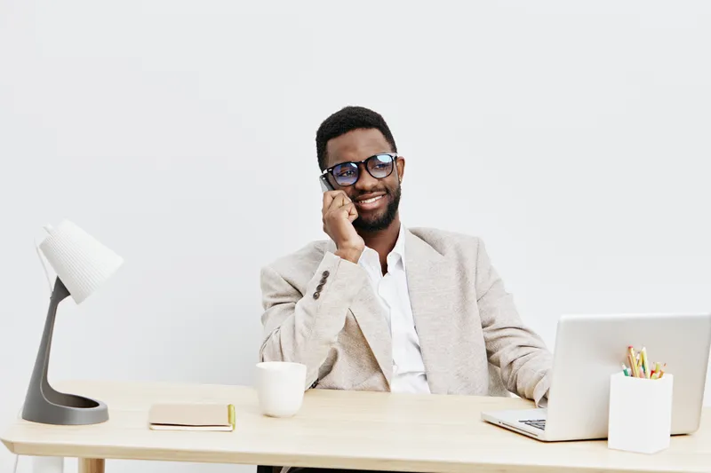 Smiling man in glasses wearing beige blazer working at modern office desk with laptop, coffee mug, and notebook. Confident, professional, diverse workplace, casual style, bright background, business l