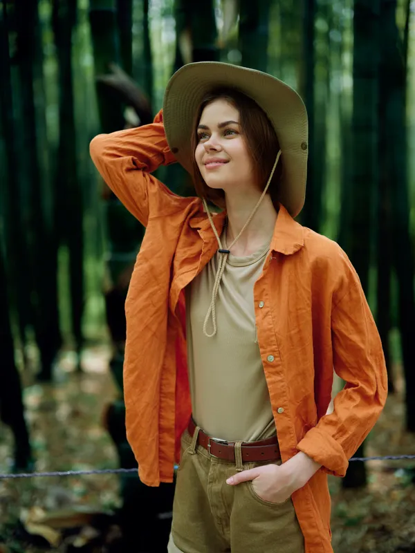 Woman in forest wearing wide brim hat and orange shirt, smiling portrait among tall trees. Casual outdoor outfit, hand on hat and pocket, relaxed nature hike and autumn mood.