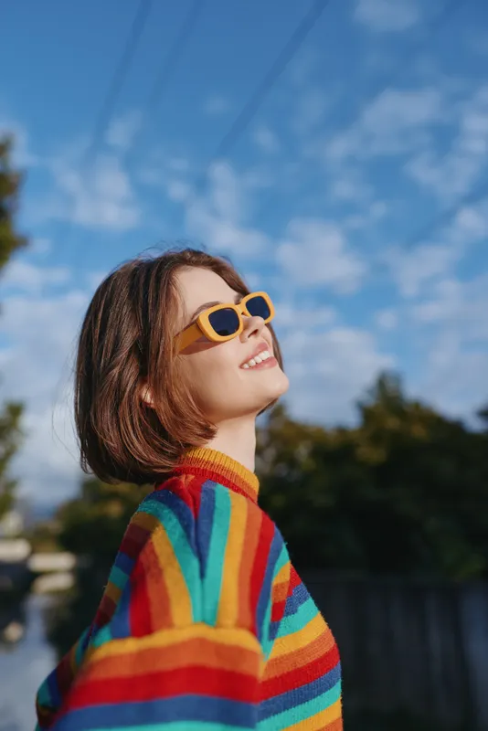 Young woman in a rainbow sweater and yellow sunglasses smiling outdoors under a blue sky, short bob haircut and casual outfit on a suburban street, joyful lifestyle.