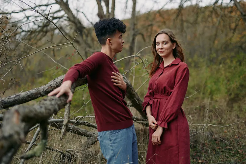 A young couple in natural autumn woods sharing an authentic, candid moment, woman in a red dress and man leaning on a fallen tree, warm tones and subtle emotion conveying genuine connection.
