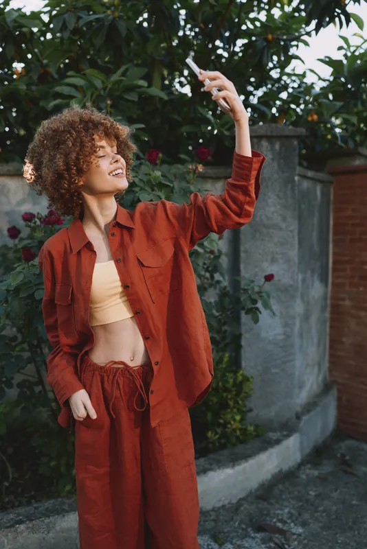 Young woman with curly hair wears a rust suit and crop top, taking a confident selfie outdoors as she raises her arm, smiling against a leafy garden wall