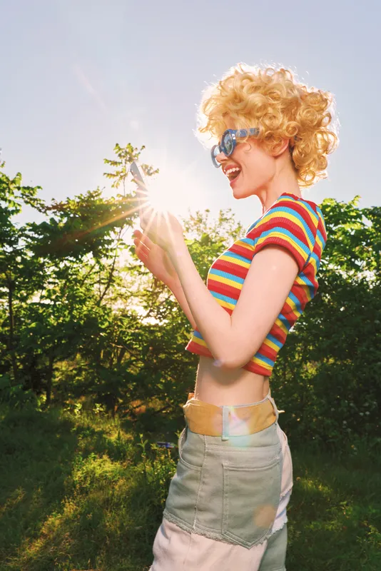 A young woman with curly blonde hair and sunglasses stands outdoors in a sunlit park, smiling as she catches the warm light with a playful, carefree pose.