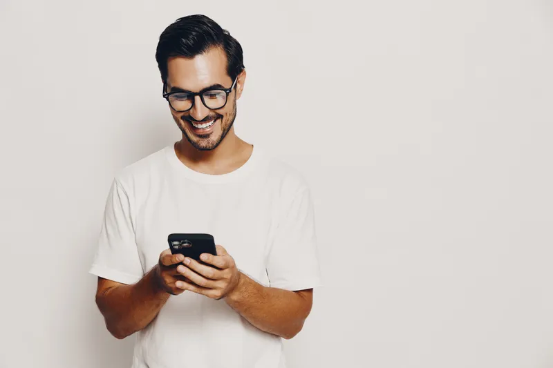 Smiling young man wearing glasses and a white t-shirt holds a smartphone while looking at the screen, isolated on plain light background. People lifestyle concept.