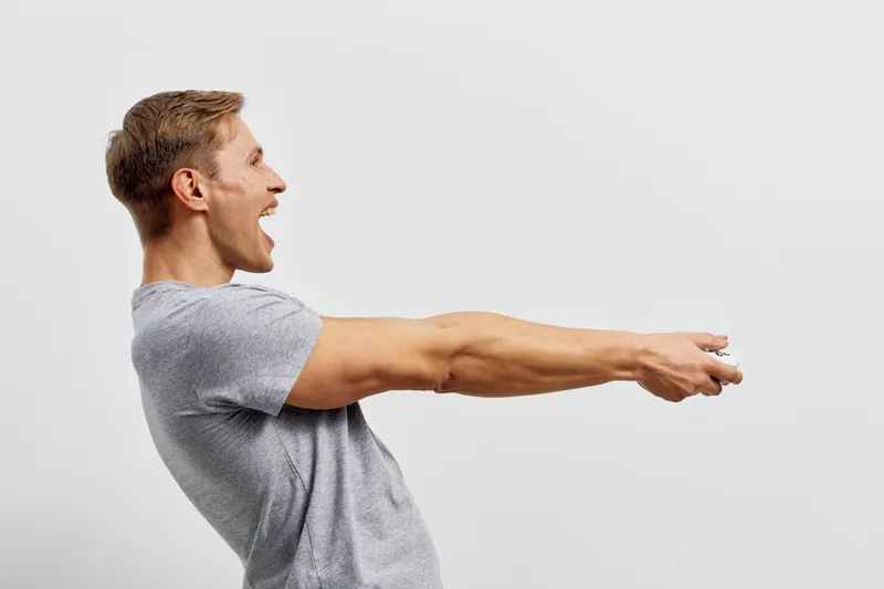 Vibrant young man with short brown hair, wearing a casual gray t-shirt, demonstrating an energetic pose with arms extended forward, expressing enthusiasm and confidence. Studio shot with plain white b