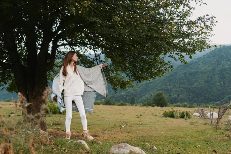 Cheerful woman outdoors, standing by a tree with a fluttering scarf, relaxed pose, sunlit landscape, natural colors, peaceful countryside scene
